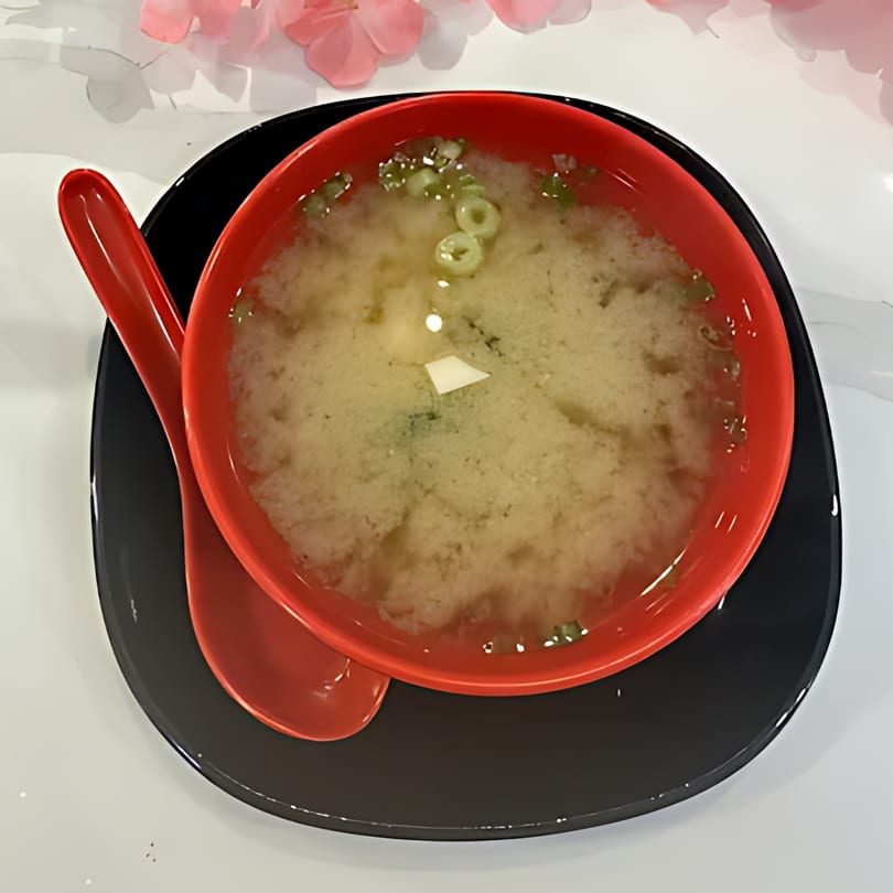 A red bowl of miso soup with green onions and tofu, served on a black plate with a red spoon.