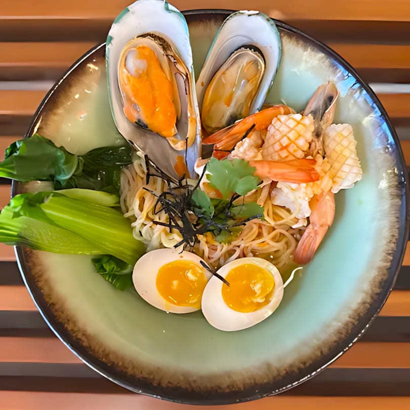 Seafood ramen with mussels, shrimp, squid, bok choy, and soft-boiled eggs in a light green bowl.