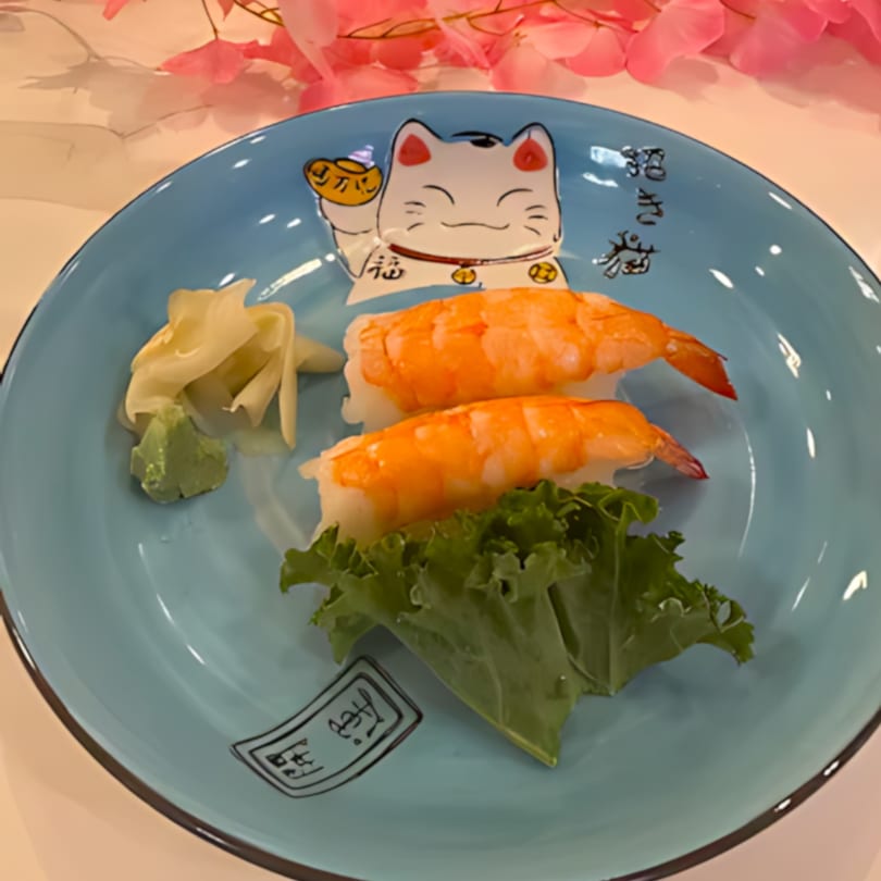 Two shrimp nigiri sushi pieces with ginger, wasabi, and lettuce on a blue Maneki-neko plate.